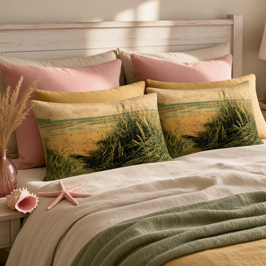 Two decorative throw pillows with a beach grass print, styled on a bed with pink and cream pillows, a linen cover, and a green throw blanket.