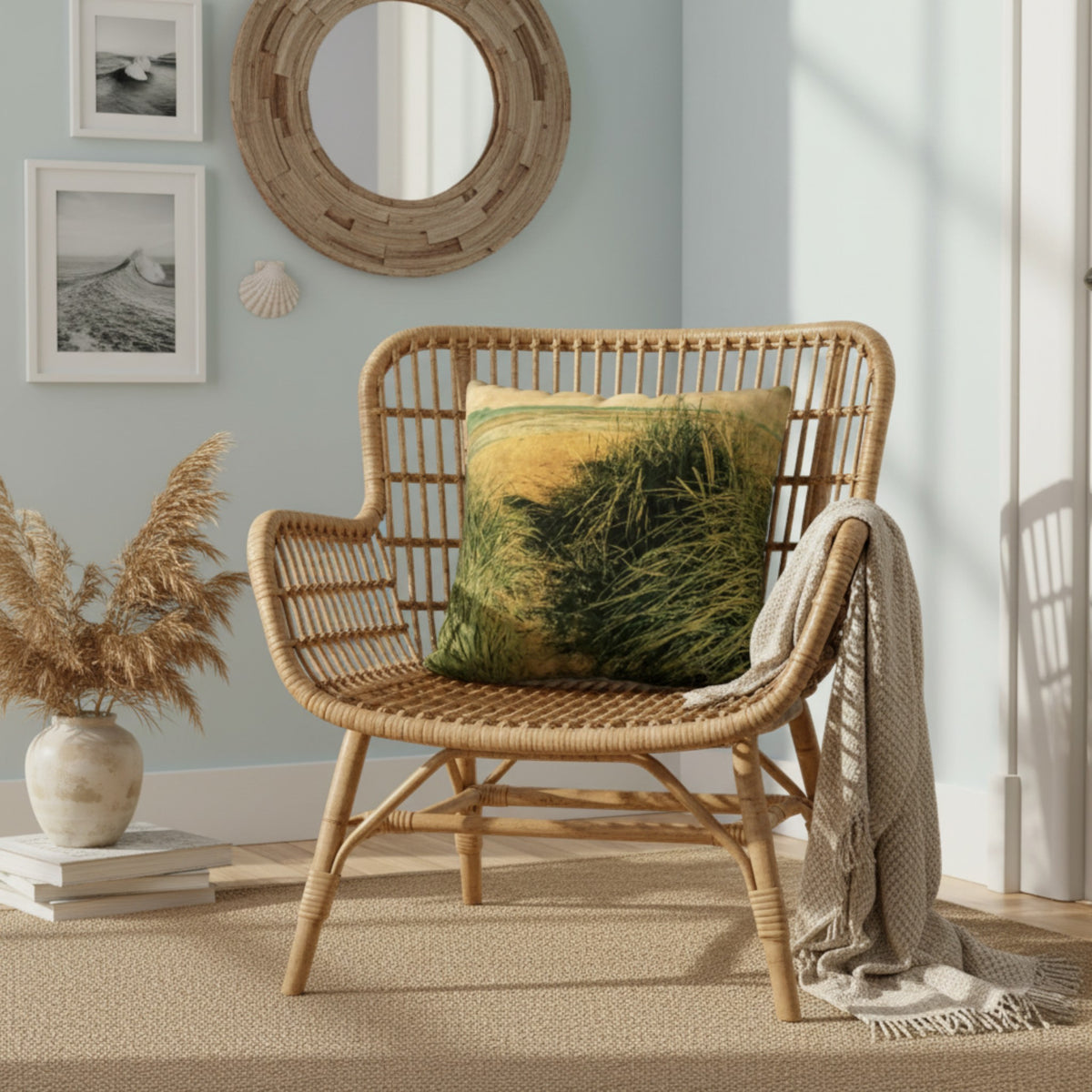 A woven rattan armchair in a styled room setting, featuring a square throw pillow with a green and yellow grassy field print and a beige knit throw blanket draped over the side.