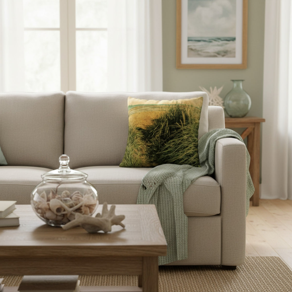 A beige upholstered sofa in a bright living room, styled with a green and yellow grass-patterned throw pillow, a light green knit throw blanket, and a wooden coffee table in front.