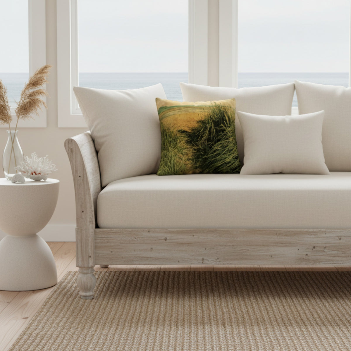 A white upholstered beach-style sofa with a distressed whitewashed wood frame, styled with cream pillows and a square throw pillow featuring a green and yellow grassy field print. The sofa is set in a bright room with a window overlooking the ocean, next to a white side table.