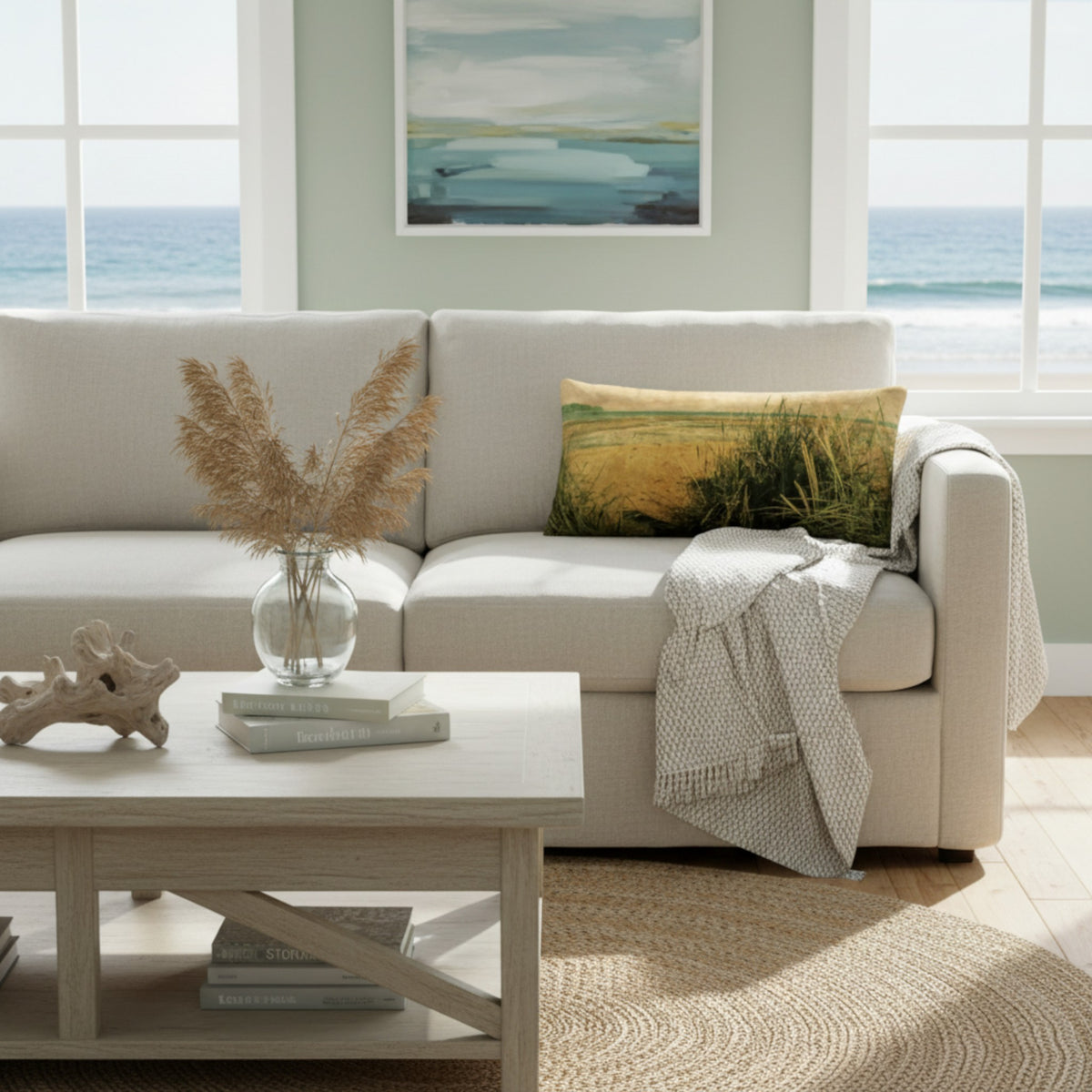 A light beige sofa in a bright coastal living room, styled with a green and yellow grass-patterned throw pillow, a white throw blanket, a glass vase with dried grass, and a light wood coffee table.