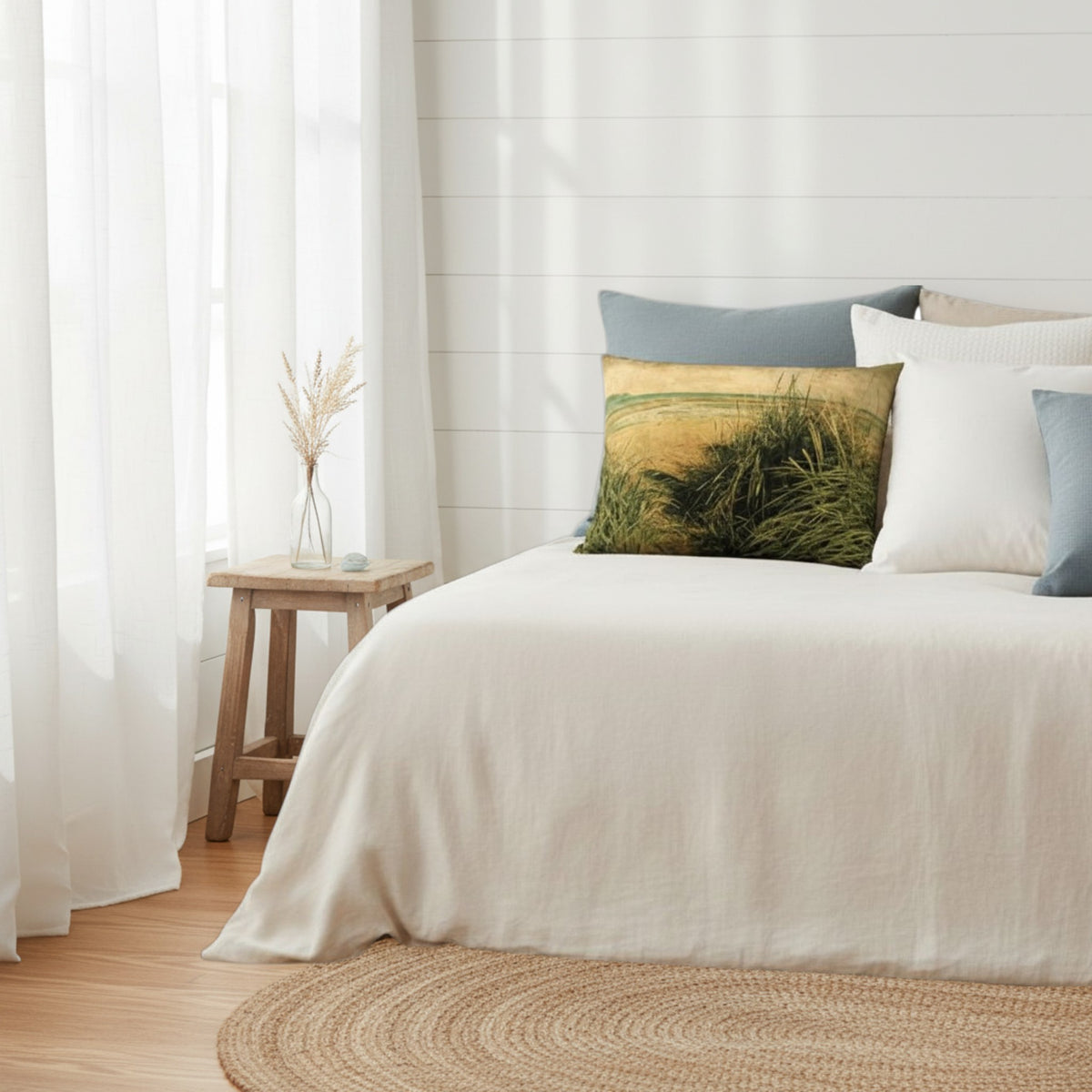 A styled bedroom scene with a bed dressed in white bedding and several decorative pillows, including a square throw pillow featuring a grassy landscape print. A small wooden side table with a vase stands beside sheer white curtains and a light wood floor.
