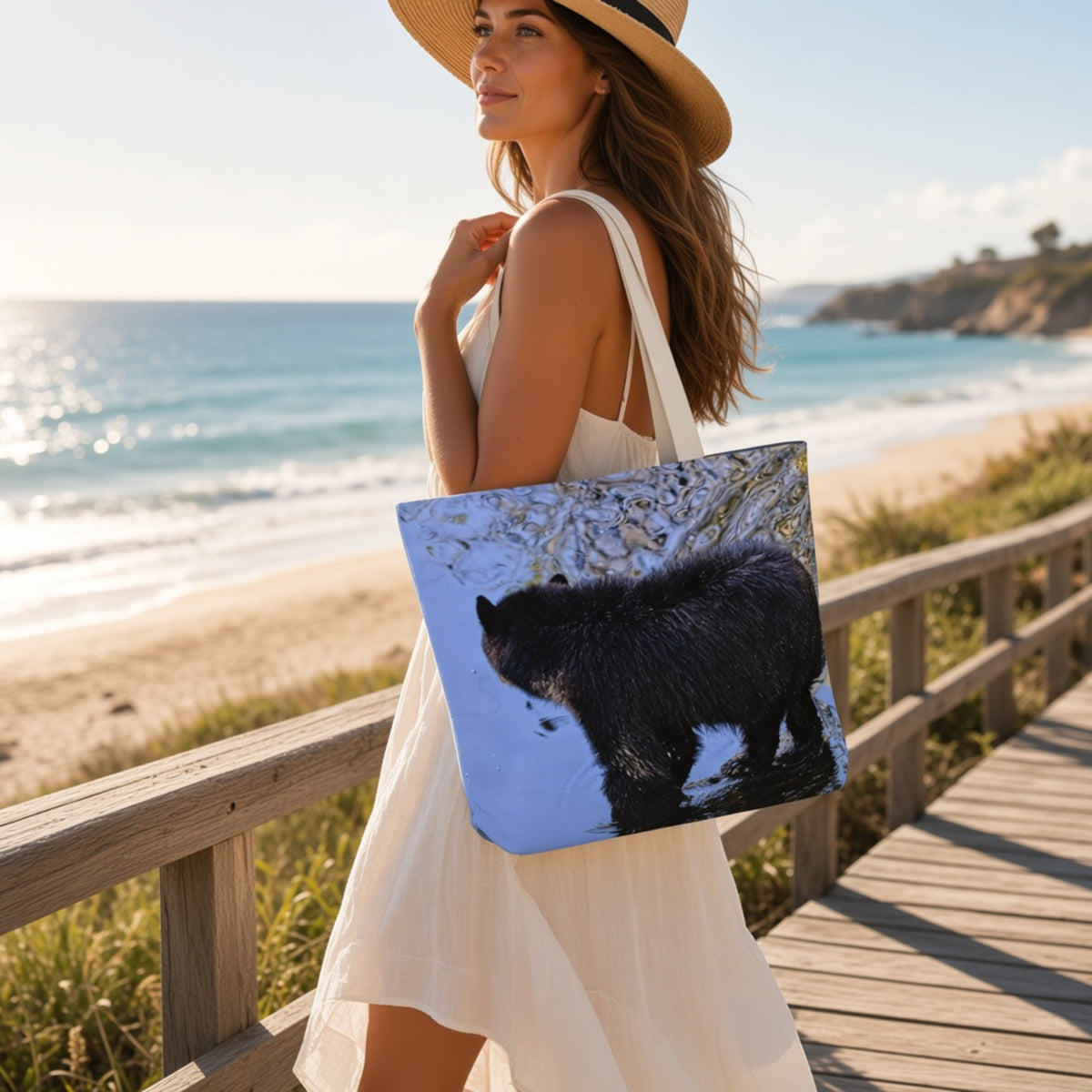 A woman in a white dress and straw hat carries a tote bag on a wooden boardwalk by the beach. The tote bag features a printed design of a black bear standing in shallow water.