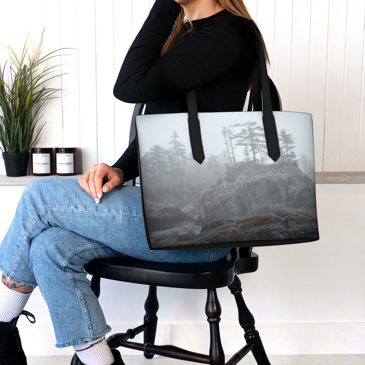 A woman sitting on a black wooden chair holds a large tote bag with a misty forest and rocky landscape print. The woman is wearing a black long-sleeve top, ripped blue jeans, white socks, and black shoes in a bright room with a white paneled wall, a potted plant, and candles.