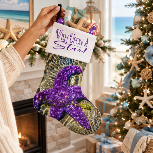 A person holds a Christmas stocking decorated with a purple glitter starfish and a starry night sky print, featuring a white cuff that reads 'Wish Upon A Star!'. The stocking is shown in a cozy, decorated holiday home setting next to a lit Christmas tree.