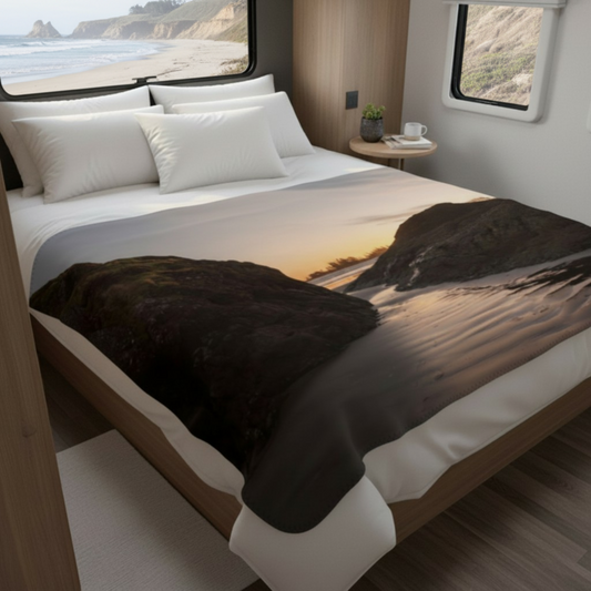 Van Isle Goddess Distant Sunset Tofino Coastal Blanket, featuring a serene beach sunset scene with rocky outcrops and gentle waves. Made of soft polyester.