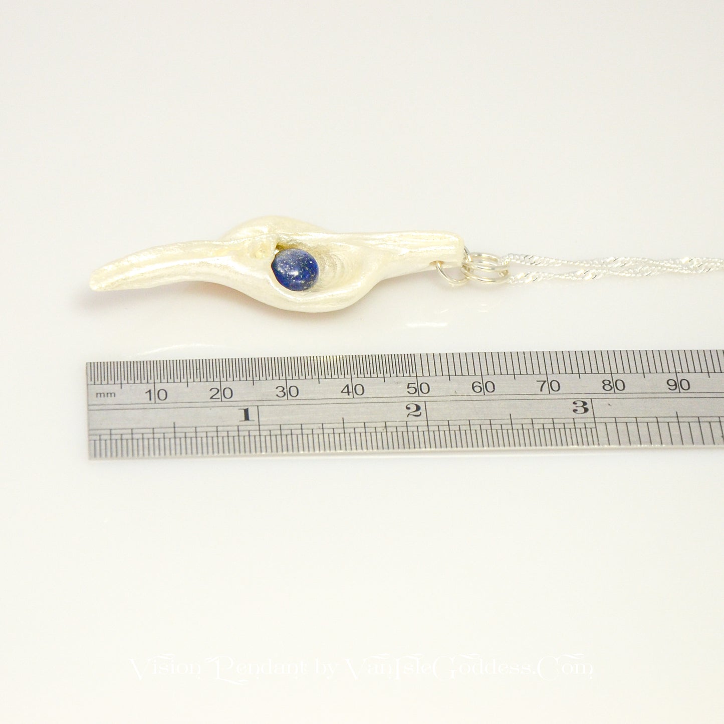 Vision is a natural seashell pendant with a Lapis Lazuli gemstone.  The pendant lays along side a ruler so the viewer can see the length of the pendant.