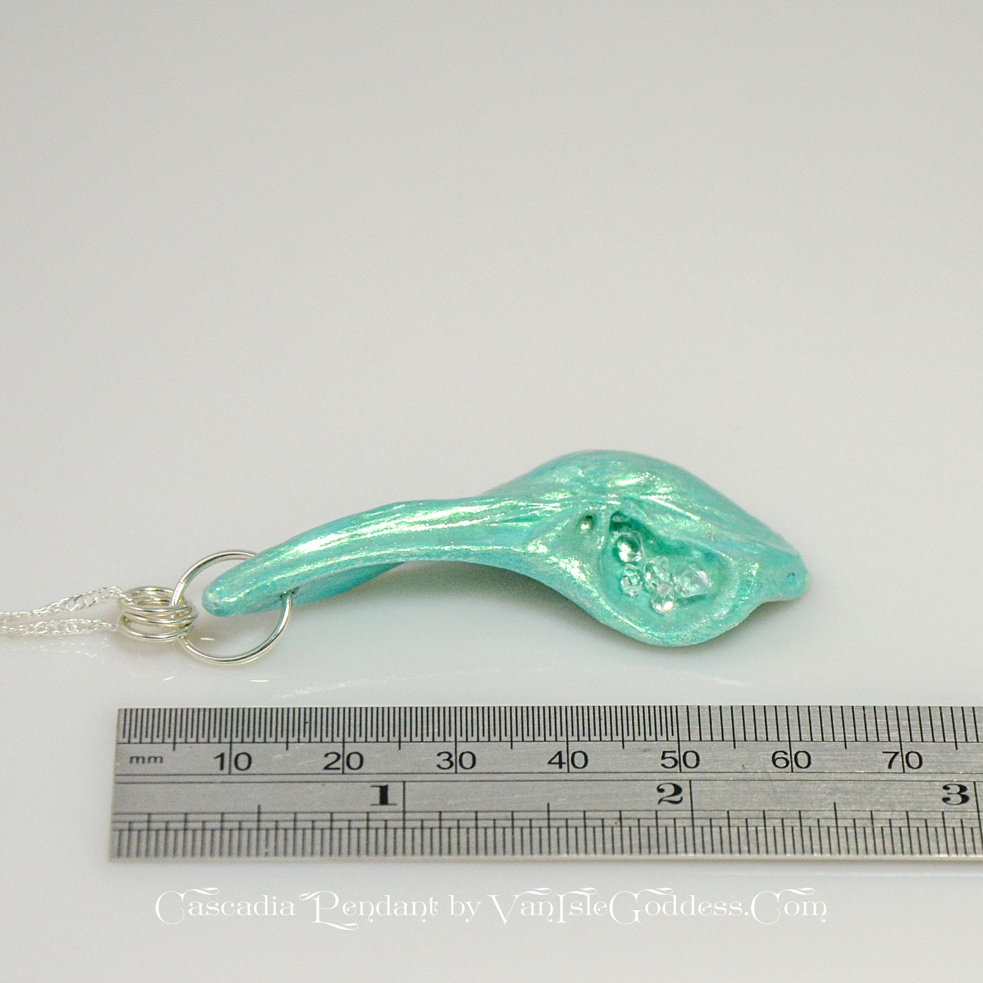 The Cascadia Pendant created with a natural seashell from the beaches of Vancouver Island.  The seashell is turquoise and has high quality herkimer diamonds. The pendant is shown on its side with a ruler so the view can see the length of the pendant. The print on the bottom of the image says: Cascadia Pendant by Van Isle Goddess dot com