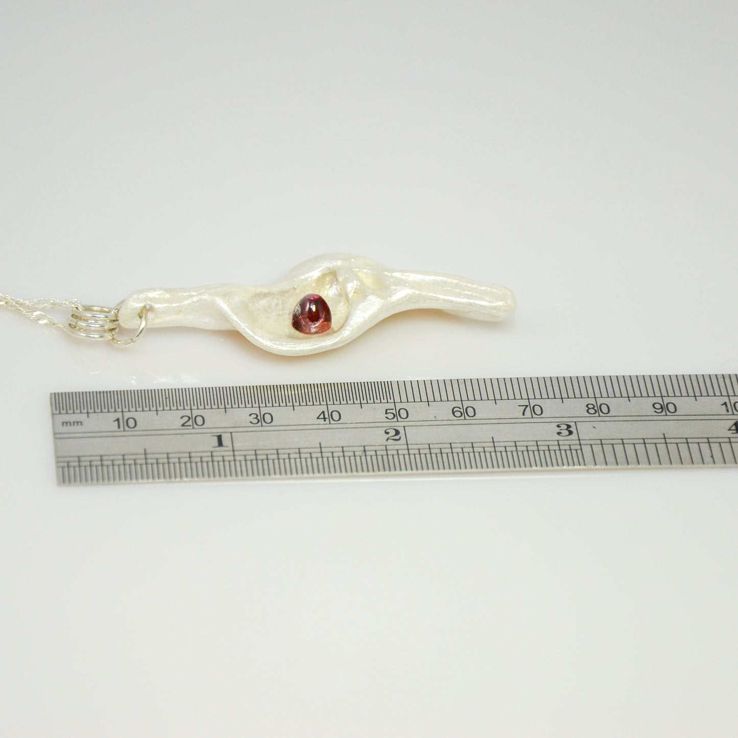 Pomegranate natural seashell pendant is adorned with a stunning Garnet gemstone. The pendant lays along side of a ruler to show the viewer the length of the pendant.